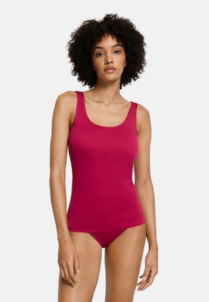 Kiharatukkainen nainen, jolla on istuva magentanvärinen toppi ja siihen sopivat alushousut, seisomassa yksivärisen valkoisen taustan edessä.