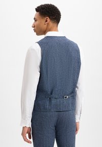 Blau gestreifter Weste mit tailliertem Schnitt, aus strukturiertem Stoff, mit einem Rückenschnallen-Detail und getragen über einem weißen Hemd.