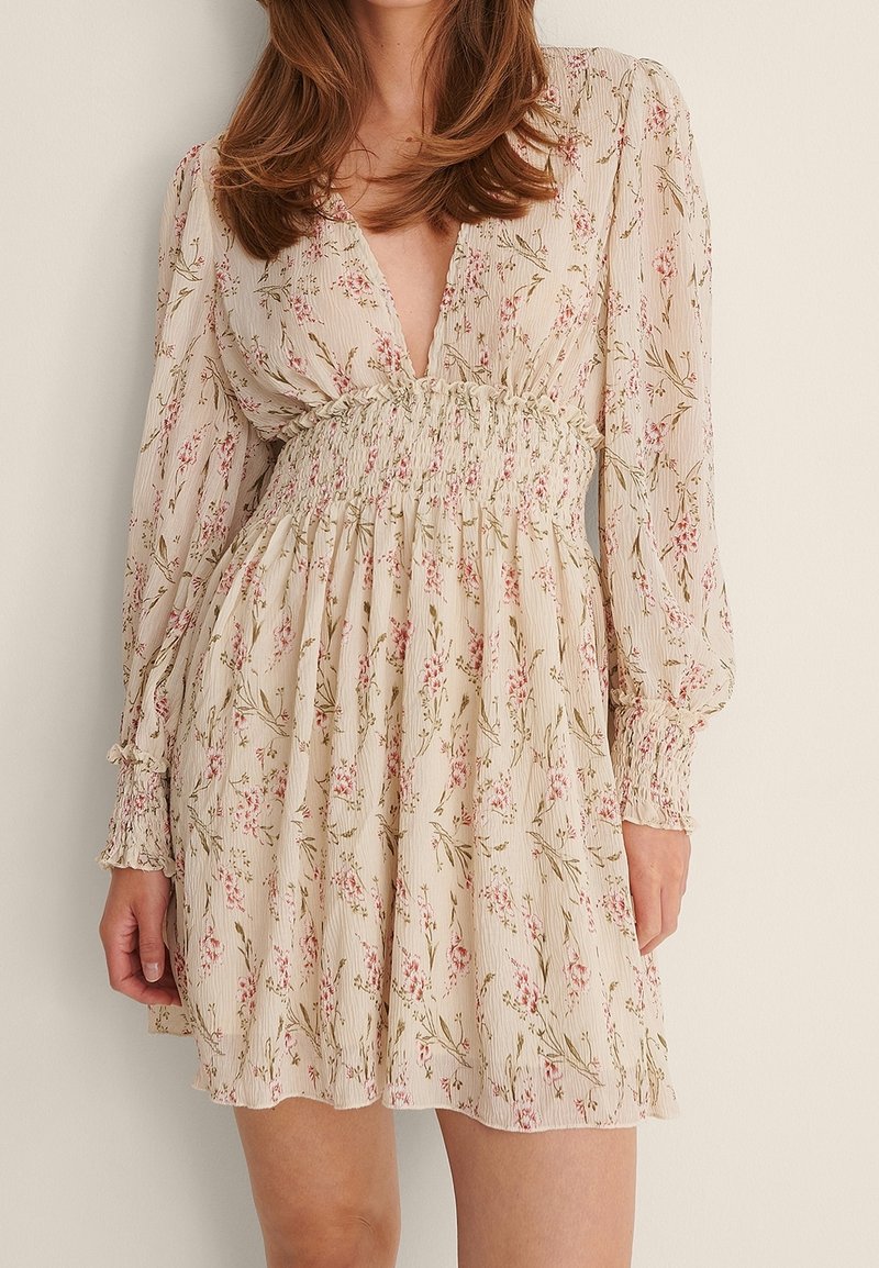 Femme portant une robe beige à motifs floraux avec des manches longues, un décolleté en V profond et une taille froncée, se tenant devant un fond neutre.