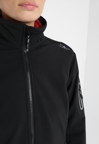 Personne portant une veste noire zippée avec un col montant et un logo sur l'épaule, doublure intérieure rouge visible au niveau du cou.