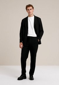 Melnas uzvalka žaketes un bikses, uzvilktas uz baltas T-krekla. Iekļauj pieguļošu dizainu, gludu tekstūru un spīdīgas melnas kurpes. Vienkāršs fons.