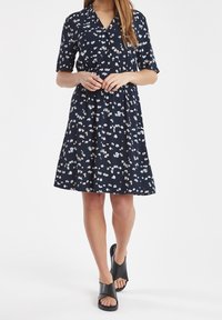 Kvinna i en marinblå knälång klänning med vit blommönster och svarta sandaler med öppen tå, stående mot en vit bakgrund.