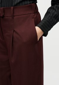 Άτομο που φοράει βερμούδα μπορντό με το χέρι στην πλαϊνή τσέπη και μαύρο πουκάμισο με λεπτές ρίγες και μακριά μανίκια.