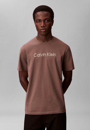Tânăr cu mâinile la spate, purtând un tricou maro Calvin Klein, pe un fundal gri simplu.