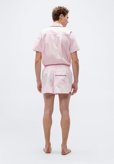 Άνδρας μοντέλο όρθιος ξυπόλητος, γυρισμένος προς τα πίσω, φοράει ανοιχτό ροζ κοντομάνικο μπλουζάκι και αντίστοιχα σορτς με πίσω τσέπη και σκούρες λεπτομέρειες.