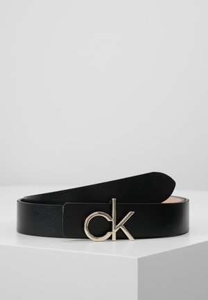 Czarny skórzany pasek z pozłacaną klamrą z logo "ck", prezentowany zrolowany na białej powierzchni na tle jednolitego jasnoszarego tła.