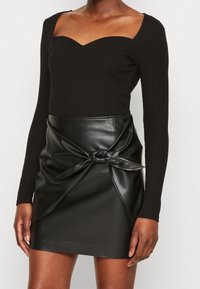 Schwarzes, geripptes Langarmoberteil mit herzförmigem Ausschnitt, kombiniert mit einem schwarzen Minirock aus Kunstleder mit einer Bindung an der Taille.