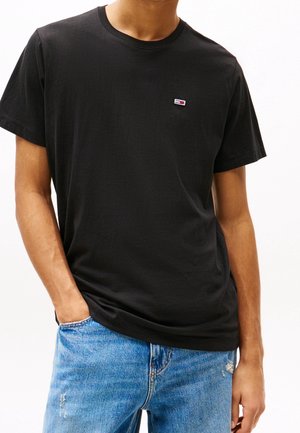Mężczyzna noszący prosty czarny t-shirt z krótkim rękawem z małym logo na piersi i niebieskie przetarte jeansy, jedna ręka w kieszeni.