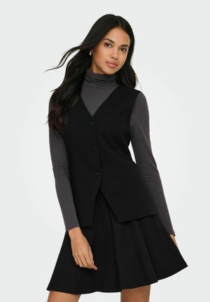 Femeie cu păr lung și închis la culoare, purtând un vestă neagră cu nasturi peste un helancă gri și o fustă neagră asortată, pozând pe un fundal alb.