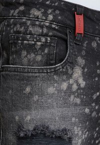 Czarne dżinsy z materiału denim w stylu przetartym, z plamkowanym wzorem, postrzępionym dołem i czerwoną metką z tkaniny na pasku.