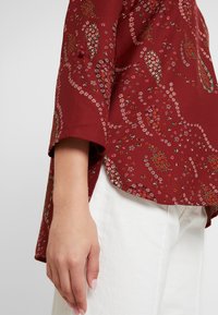 Blouse à motifs floraux bordeaux avec des manches courtes, dotée d'un ourlet arrondi et de délicates accents de fleurs blanches et vertes. Portée avec un pantalon blanc.