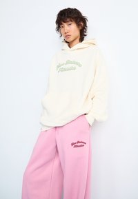 Sweatshirt à capuche crème avec logo "New Balance Athletics" vert, associé à un pantalon de jogging rose arborant le même logo en rouge. Les pantalons de jogging possèdent des poches.