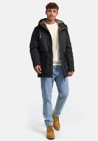 Mørkeblå isoleret jakke med en hætte i brunt pels, brune accenter, flere lommer, båret over en cremefarvet sweater og lyseblå jeans.