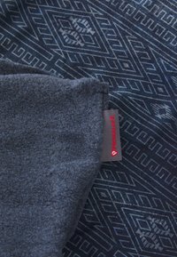 Mørkeblå fleece tæppe med geometriske mønstre, tekstureret overflade og et Primaloft mærke. Har en blød, varm følelses med detaljerede designs.