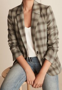 Marynarka w kratę w odcieniach szarości i zieleni, z klapami i zapięciem na jeden guzik, w połączeniu z białą bluzką i niebieskimi dżinsami.