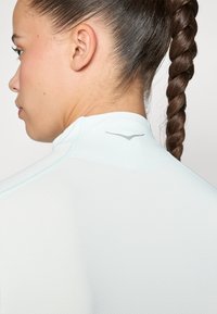 Lys blå tettsittende topp med et teksturert mønster, høy krage og en liten sølv logo aksent bak på nakken.