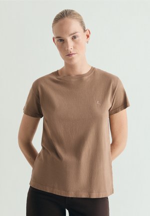 Kvinde med blondt hår bundet tilbage iført en ensfarvet brun kortærmet t-shirt og sorte bukser, stående mod en lysegrå baggrund.