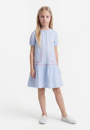 Niña joven de pie con un vestido azul claro de manga corta con bolsillos delanteros y zapatos blancos sin cordones sobre un fondo blanco liso.