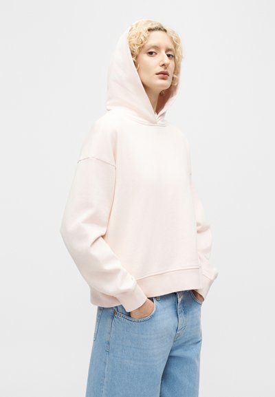 Άτομο με σγουρά ξανθά μαλλιά, ντυμένο με ανοιχτόροζ φούτερ με κουκούλα και μπλε τζιν, με τα χέρια στις τσέπες, να στέκεται μπροστά σε απλό φόντο.