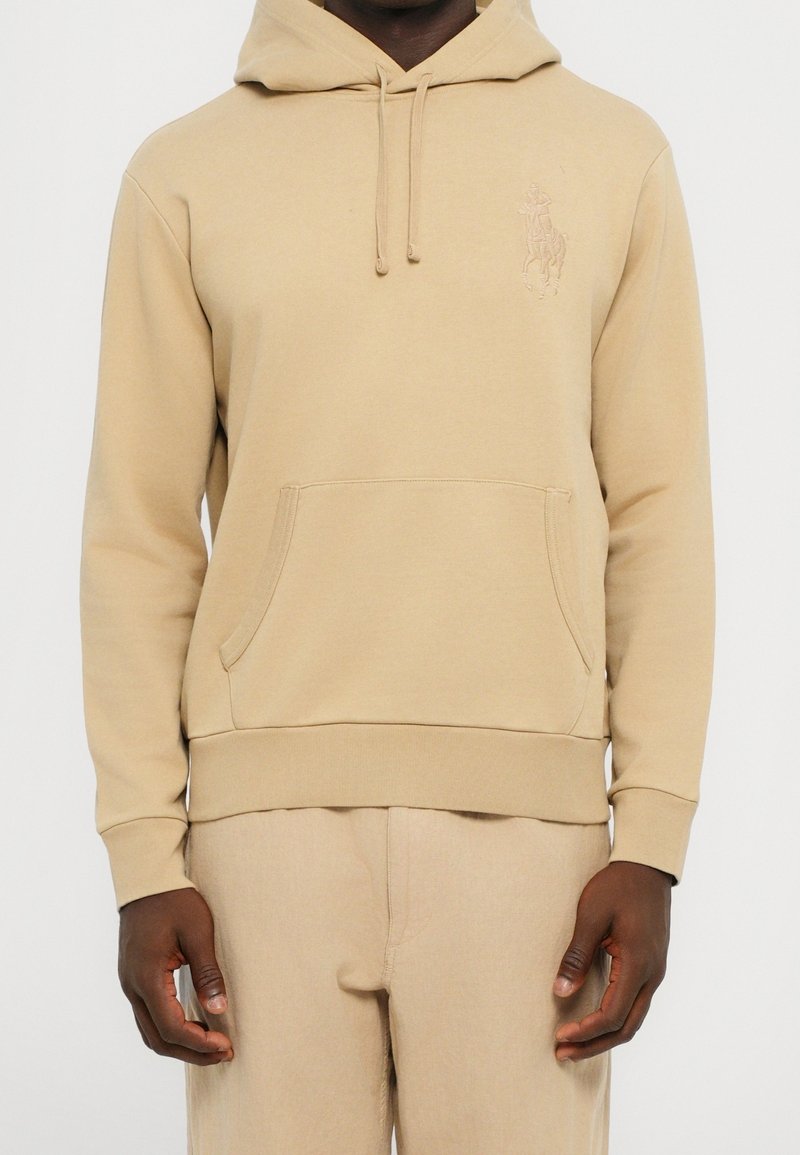 Homme portant un sweat à capuche beige avec poche ventrale avant et pantalon beige assorti, debout devant un fond neutre.