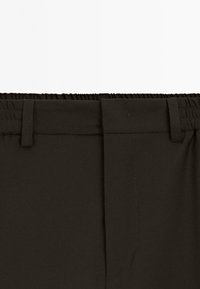 Mørkebrune bukser med elastisk talje, flad forside, bæltestropper og minimalistisk design. Glat tekstur, ingen synlige mønstre.