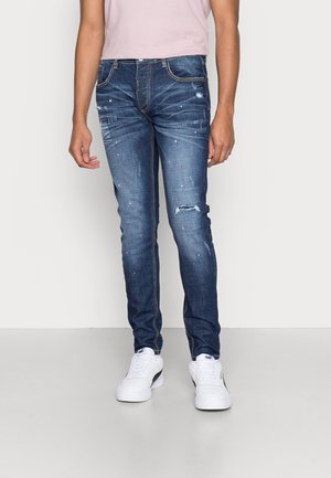 Personne portant un jean bleu slim déchiré avec des éclaboussures de peinture, une chemise rose clair et des baskets blanches debout sur un sol uni.