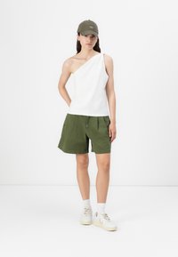Un top alb asimetric, pantaloni scurți verzi măslinii și o caciulă verde, completate cu adidași albi și șosete până la gleznă, pe un fundal simplu.