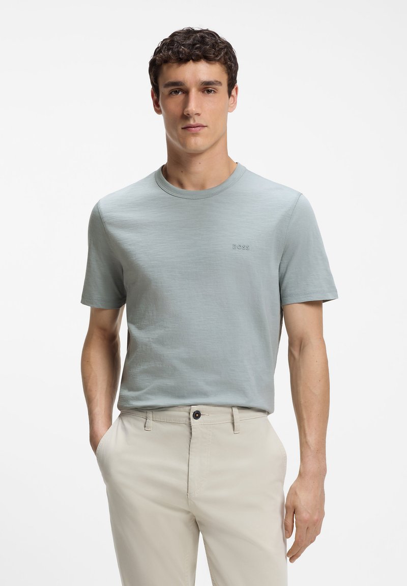 Jeune homme aux cheveux courts et bouclés portant un t-shirt bleu clair à manches courtes et un pantalon beige, debout avec une main dans la poche.