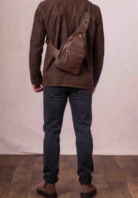 Brown leather sling bag with a textured surface, adjustable strap, and front zip pocket, worn over a brown suede jacket and dark jeans.
