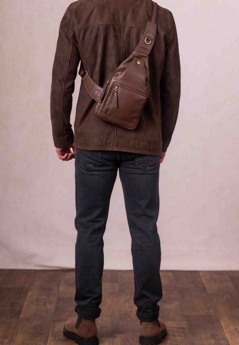 Brown leather sling bag with a textured surface, adjustable strap, and front zip pocket, worn over a brown suede jacket and dark jeans.