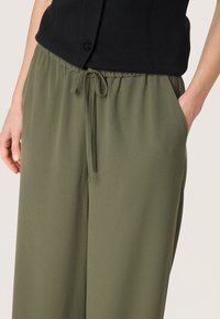 Olive green trousers with an elastic waistband, adjustable drawstring, and side pockets; smooth, lightweight fabric with a relaxed fit.