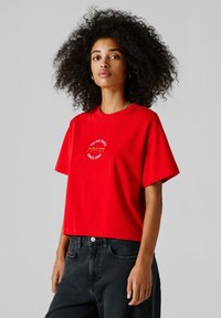 Vrouw met krullend haar, gekleed in een rood cropped T-shirt met witte tekst en logo, gecombineerd met high-waisted zwarte jeans, staand tegen een grijze achtergrond.