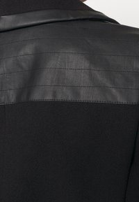 Tela negra texturizada con un panel de cuero suave en los hombros que presenta costuras horizontales. El diseño general es estructurado y moderno.