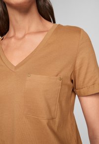 Brun kortärmad t-shirt med V-ringning i mjukt material. Har en bröstficka med mässingsknappdetaljer. Slät textur med en avslappnad passform.