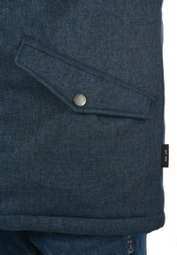 Donkerblauwe jas van een gestructureerde stof, met een klepzak met een metalen drukknoop en een klein label met de tekst "EST 1993" aan de zijkant.