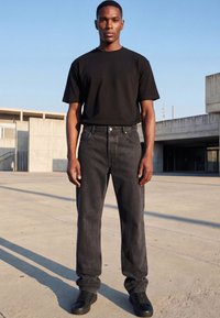 Tânăr stând în aer liber, purtând un tricou negru, blugi închiși la culoare și adidași negri pe un fundal urban din beton.