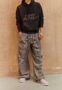 Black hoodie with multicolored "Nike" logo, grey cargo pants with multiple pockets, and purple/white checkered slip-on shoes.