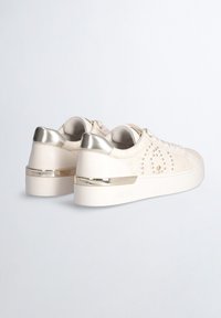 Hvide sneakers med prægede mønstre, metalliske detaljer og polstret krave. Har en chunky sål med gulddetaljer ved hælen.