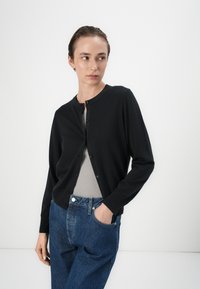 Cardigan nero con scollatura rotonda, chiusura frontale con bottoni e maniche lunghe, abbinato a un top grigio a coste e jeans blu a vita alta.