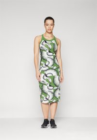 Vestido sem mangas com uma base verde, apresentando um padrão de pássaros preto e branco, design ajustado e bainha de comprimento médio. Combinado com ténis pretos.