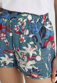 Korte broek met bloemmotief in turquoise stof en een elastische tailleband. Voorzien van witte, rode en blauwe bloemen met groene bladeren en subtiele details.