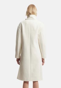 Cappotto lungo bianco con colletto alto, realizzato in un tessuto morbido e texture. Presenta spalle cadute e un design a taglio dritto.