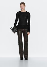 Kvinde iført en sort langærmet top, mørkebrune læderbukser, sorte støvler, der holder en sort håndtaske mod en hvid baggrund.