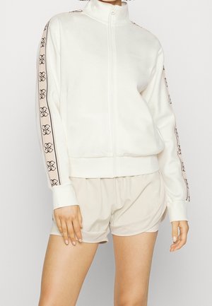 Femme portant une veste blanche zippée avec des rayures à motif sur les manches et un short de sport beige clair, debout devant un fond uni.