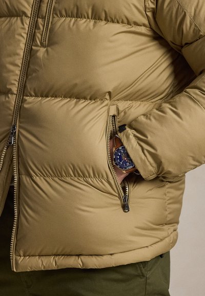 Tan puffer jacket with a zip pocket showing a hand inside, wearing a wristwatch with a blue face and brown leather strap.