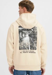 Beige Kapuzenpullover mit einem schwarz-weißen Grafikdruck auf der Rückseite, der eine abstrakte städtische Szene zeigt. Elastische Bündchen und Saum.