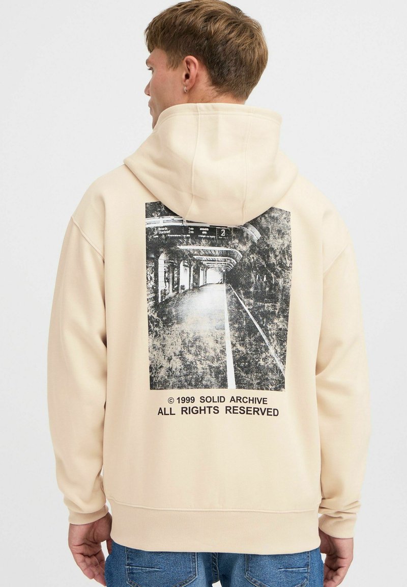 Beige Kapuzenpullover mit einem schwarz-weißen Grafikdruck auf der Rückseite, der eine abstrakte städtische Szene zeigt. Elastische Bündchen und Saum.