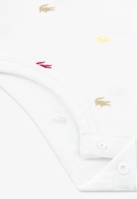 Body blanc en coton avec de petites broderies d'animaux en or, rose et beige. Comprend des fermetures à pression et un tissu doux et texturé.