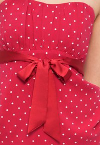Κόκκινο φόρεμα χωρίς τιράντες με λευκά πουά, διαθέτει σφηκοφωλιά σε μπροστινό μέρος και κορδέλα στη μέση. Λείο ύφασμα.