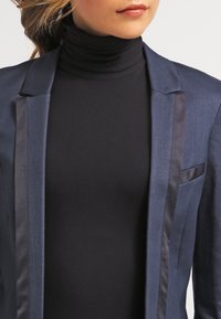 Granatowa marynarka z satynowymi klapami, o dopasowanym kroju i skrojonym fasonie; zestawiona z czarnym golfem. Skupienie na fakturze tkaniny i zorganizowanej sylwetce.
